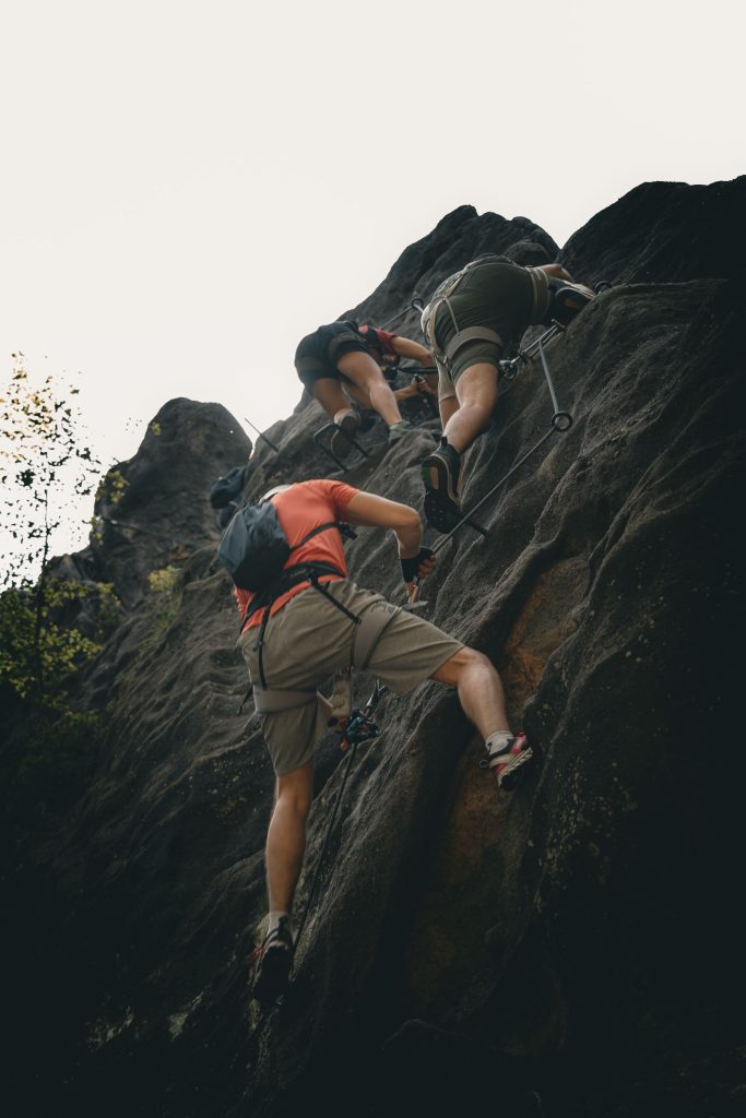 Trois pratiquants encordés progressent sur des câbles fixés à une paroi rocheuse boisée, via ferrata en moyenne montagne