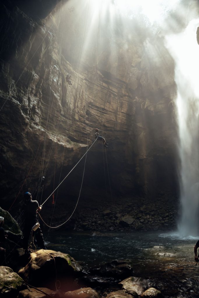 Canyonistes en rappel au fond d'une vasque encaissée, rayons de lumière traversant la cascade, illustration des tarifs du canyoning