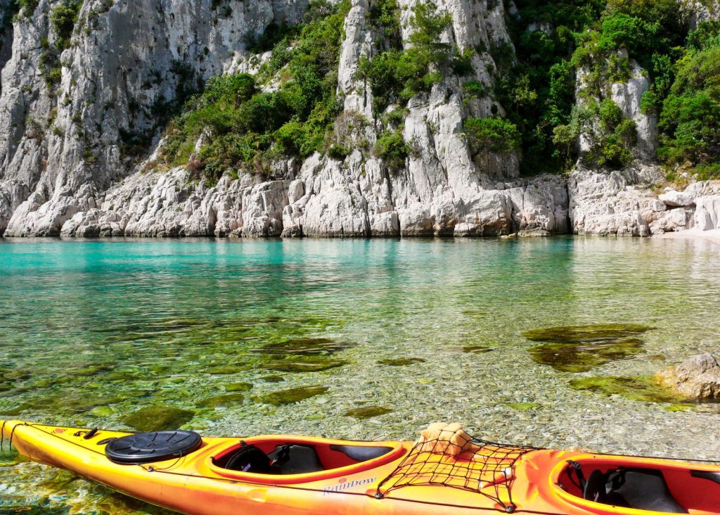 Proue d'un kayak de mer orange sur l'eau turquoise et transparente des Calanques, falaises calcaires en arrière-plan