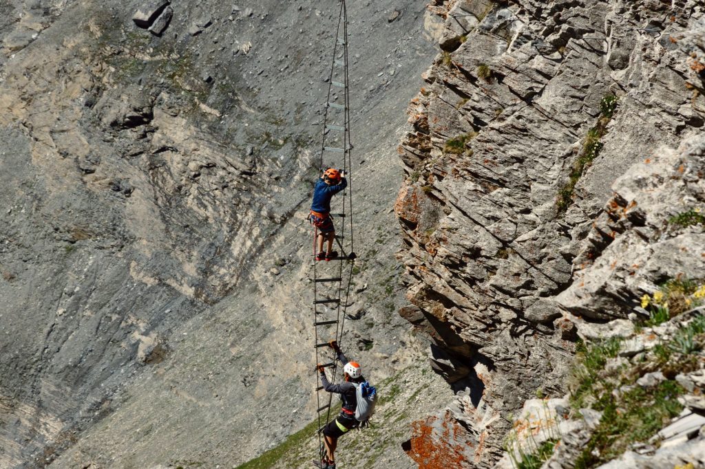 deux grimpeurs harnachés gravissant une échelle métallique sur un câble de via ferrata, paroi rocheuse