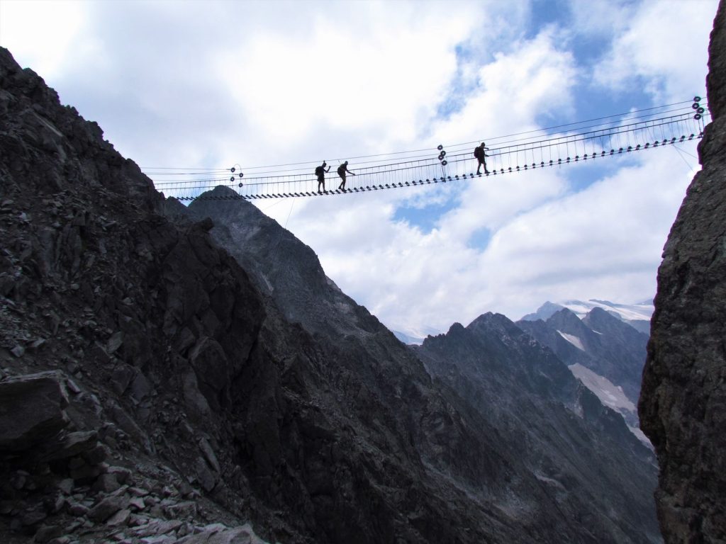 Pratiquants en silhouette traversant un pont de via ferrata suspendu au-dessus d'une vallée alpine encaissée, illustration de la via ferrata en Savoie