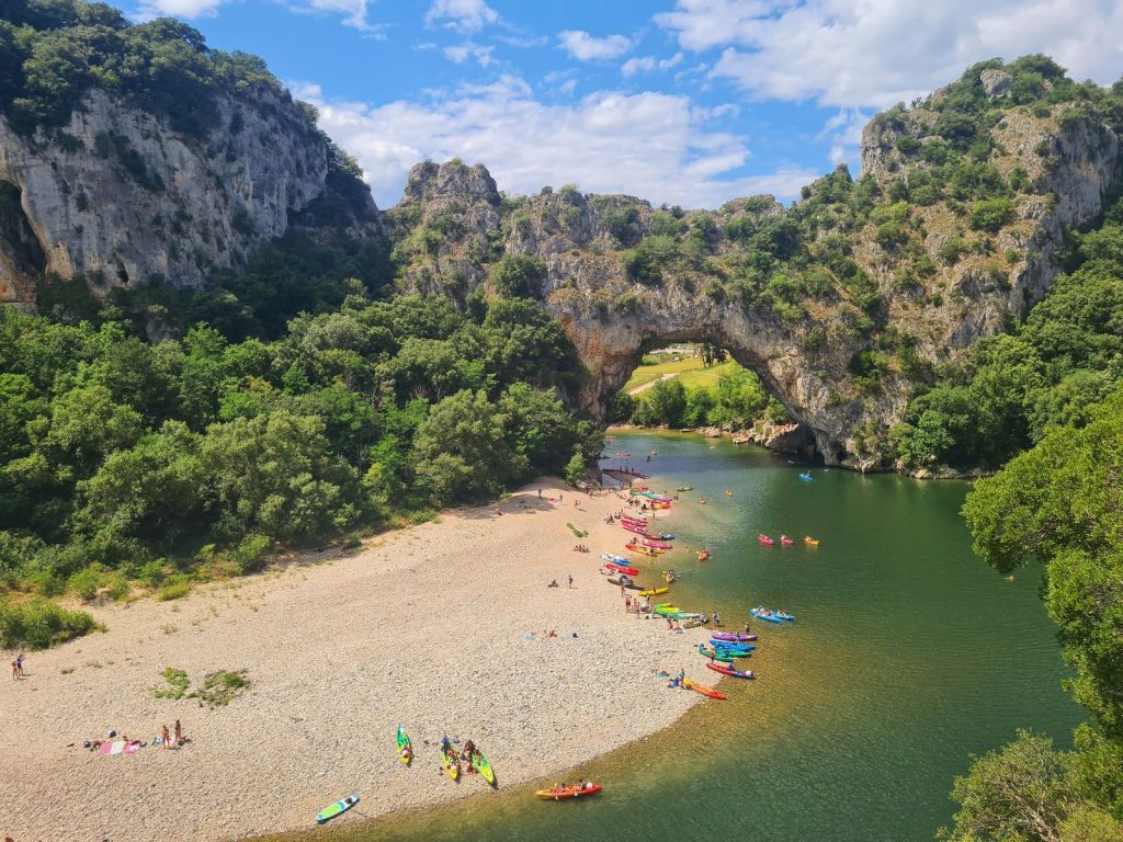Vue sur l'arche naturelle du Pont d'Arc avec des kayaks et canoës colorés sur la plage et la rivière, illustration du canoë dans les gorges de l'Ardèche