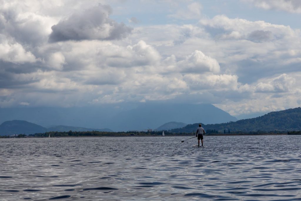 Pratiquant de paddle debout sur un grand lac alpin sous un ciel nuageux, montagnes en arrière-plan, lac du Bourget