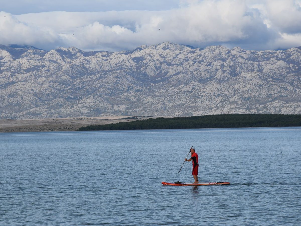 Pratiquant de paddle debout sur son stand-up paddle sur un lac calme entouré de montagnes, ciel voilé