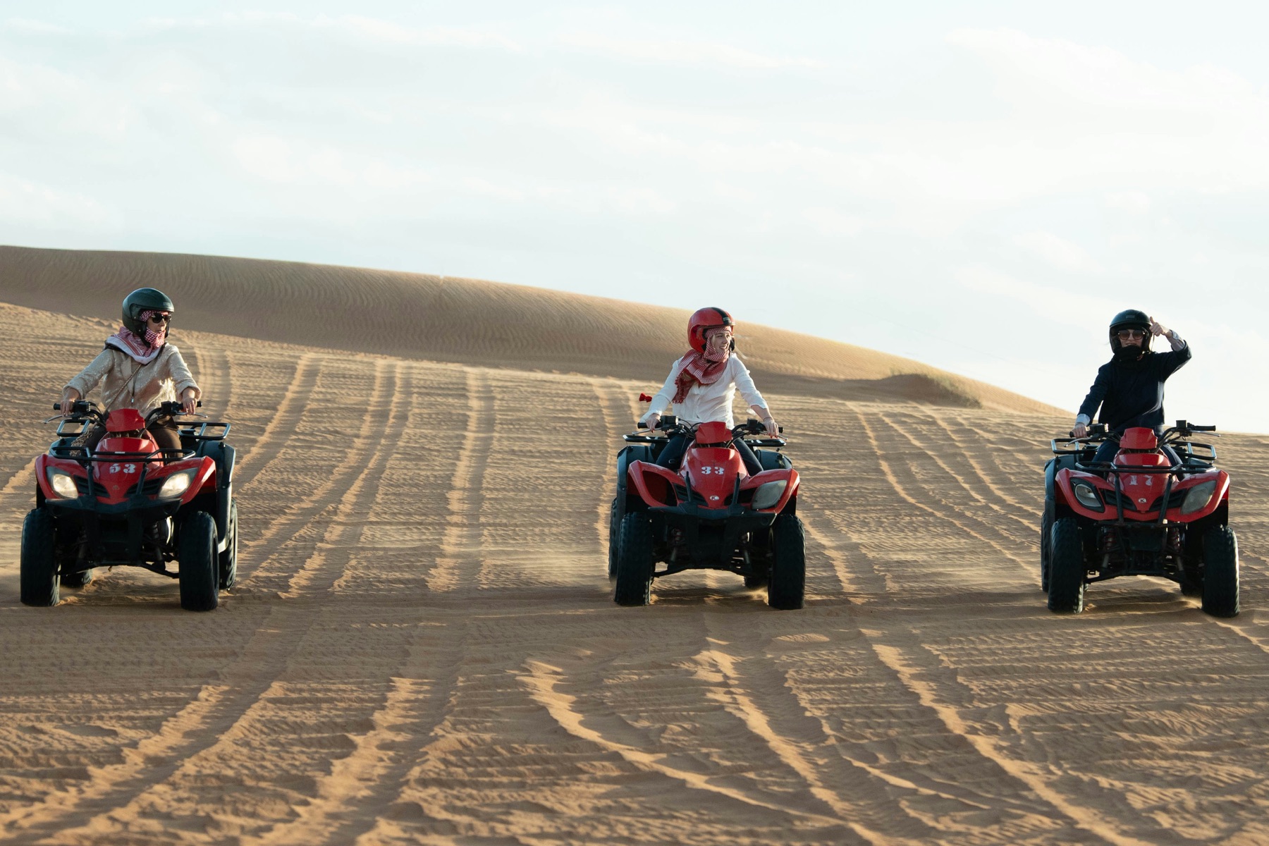 trois pilotes casqués sur des quads rouges progressant en file sur une piste dans les dunes du désert