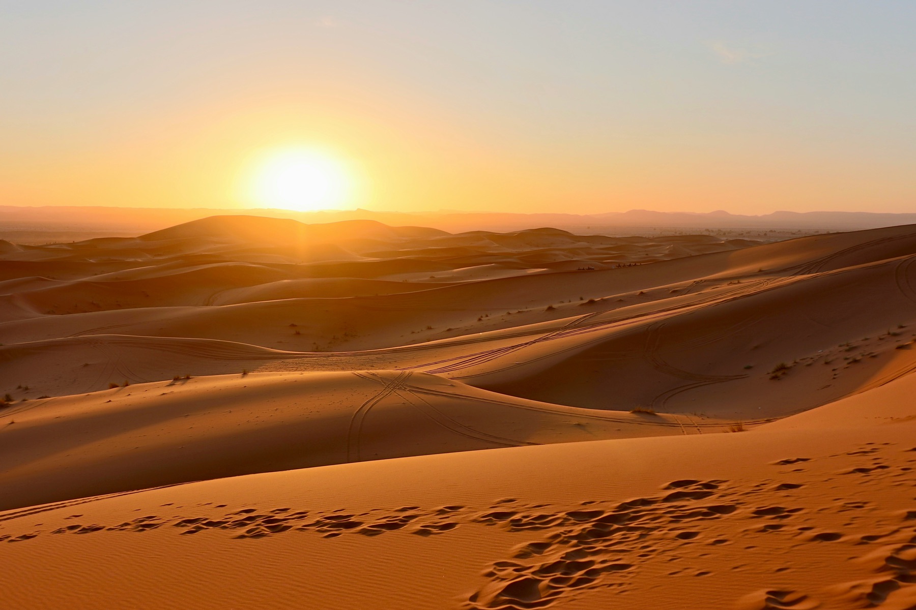 coucher de soleil orangé sur des dunes de sable avec des traces de pistes visibles au premier plan