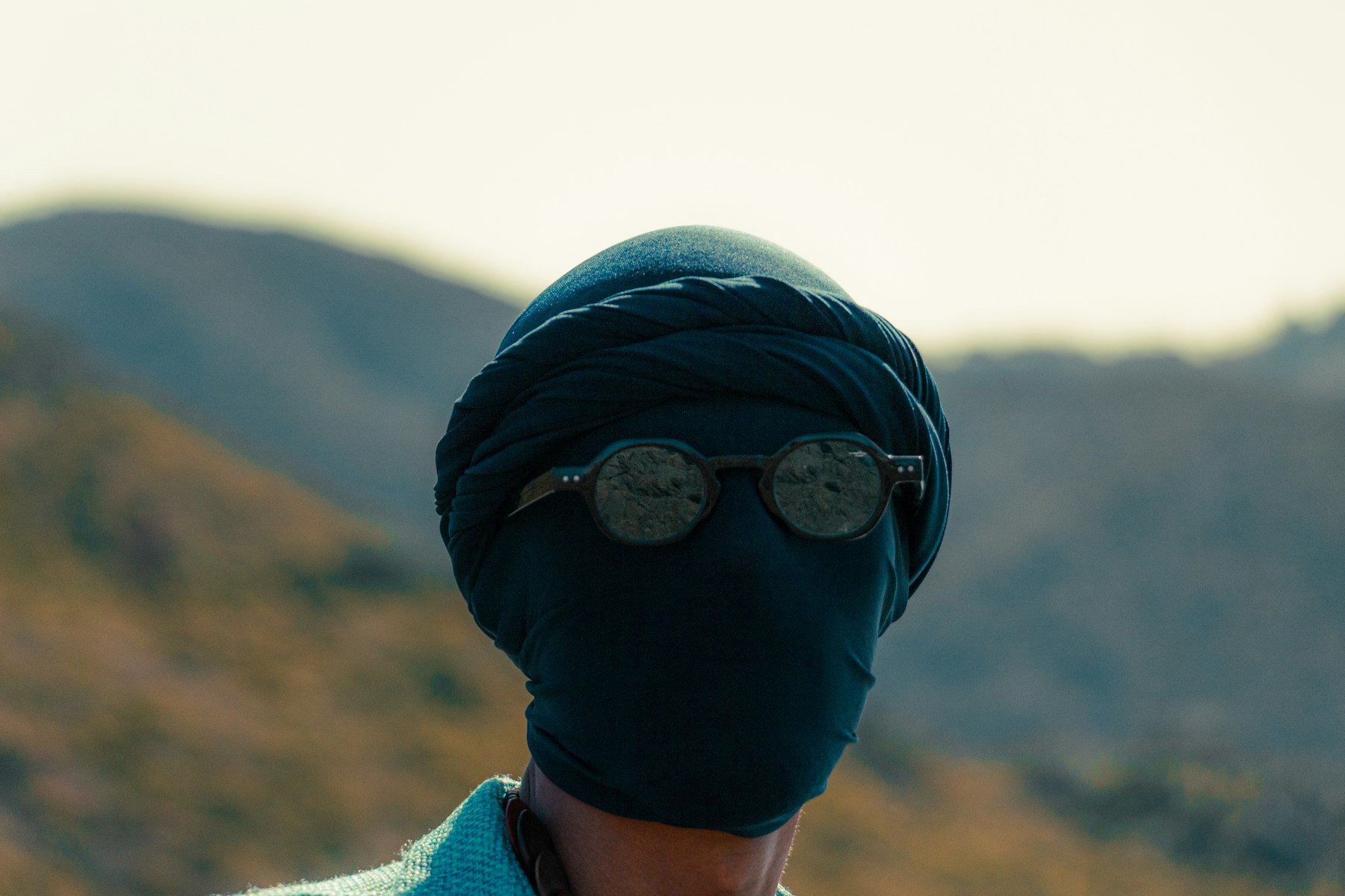visage entièrement enveloppé dans un voile sombre, lunettes rondes, collines sèches en arrière-plan